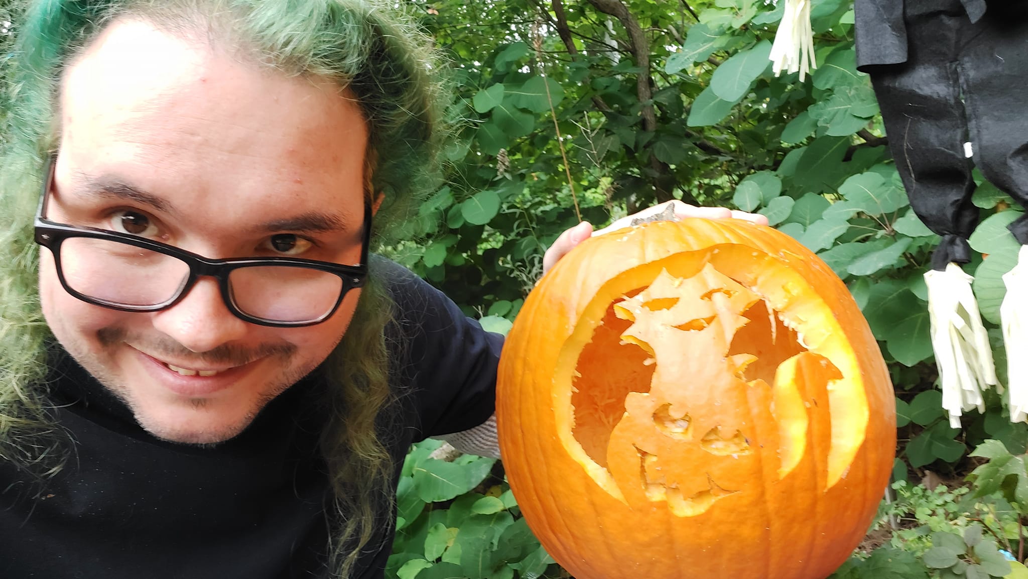 A man with long green hair and no beard holds a large punpkin with a cat poorly carved into it